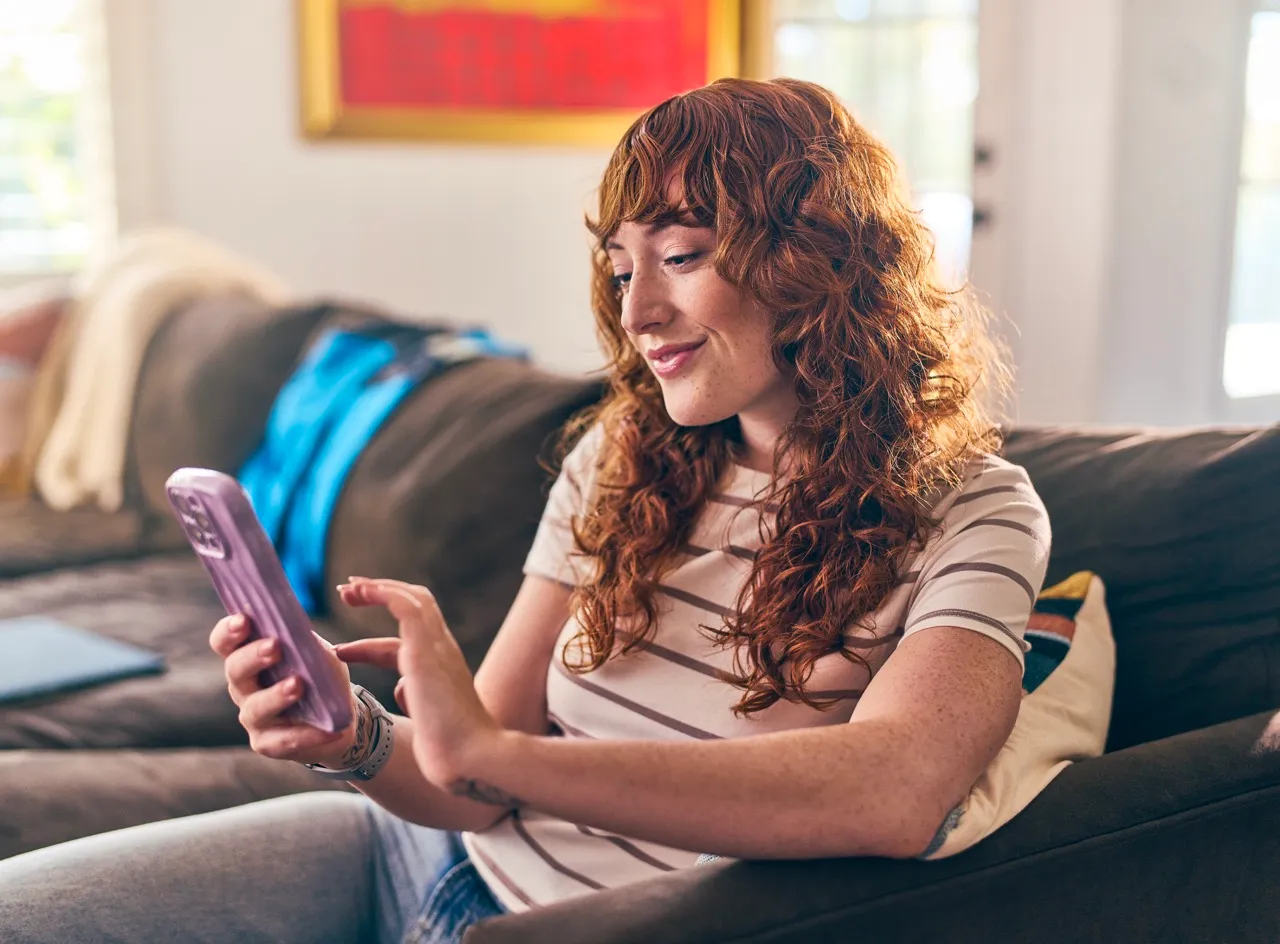 A smiling young woman sits on a couch and scrolls on her phone.
