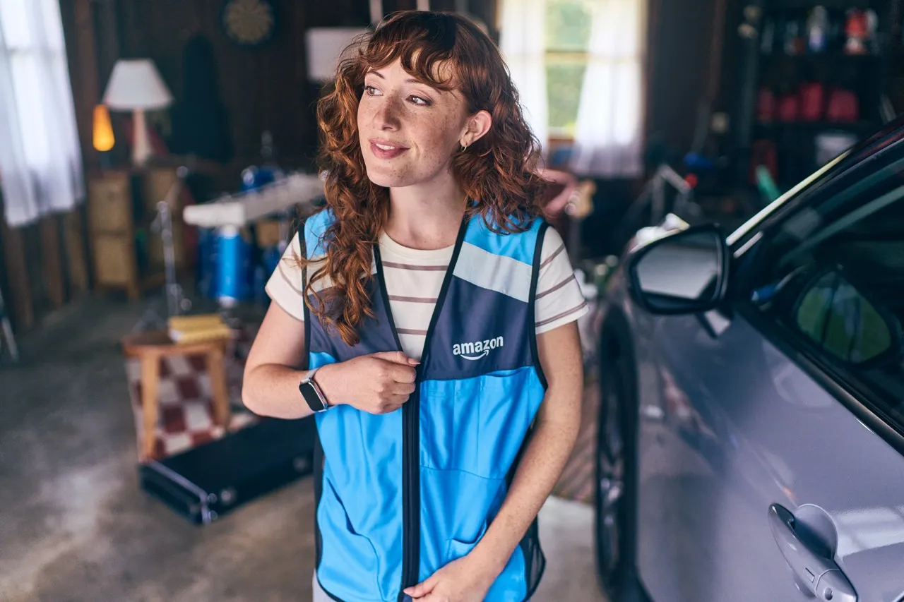 A smiling young woman stands in a garage next to a silver vehicle and zips up a blue Amazon vest.