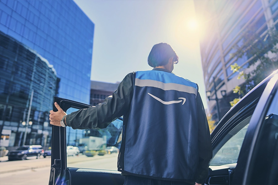A man stands behind the open door of his car on a city street between two tall glass buildings. He wears a blue Amazon vest.