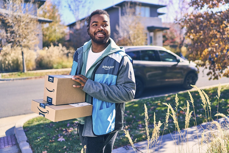 A man who wears a blue Amazon vest stands in front of an SUC and holds two packages