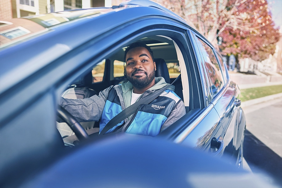 A man in a blue Amazon vest sits in the drivers seat of a car and looks out the rolled-down drivers side window