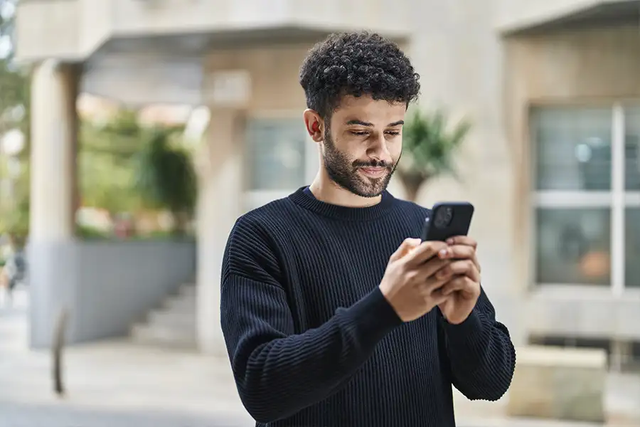 A man stands in front of an apartment building and looks down at the phone in his hands.
