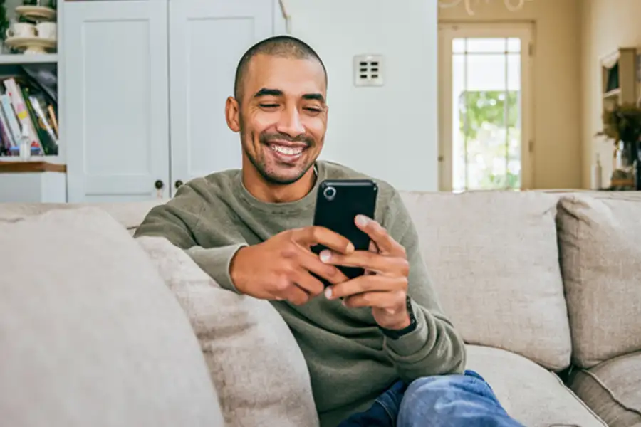 A young man sits on a couch in a living room and smiles down at a phone that he holds with both hands