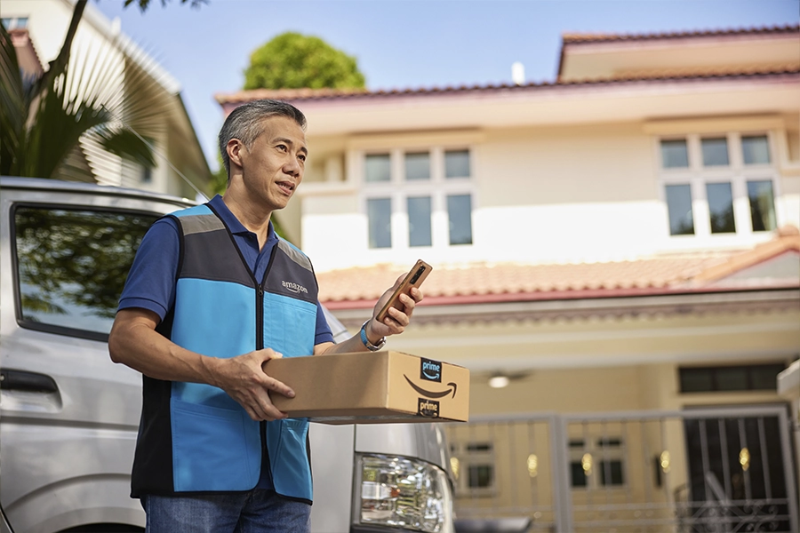 A man in a blue Amazon vest stands in front of a house and holds his phone and a package